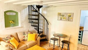 A cozy living room with a sofa, coffee table, and a small dining area with chairs. A black spiral staircase leads to an upper level. - Cotswolds Country Cottage | Boutique, Romantic Retreat for Two (Purton Stoke)