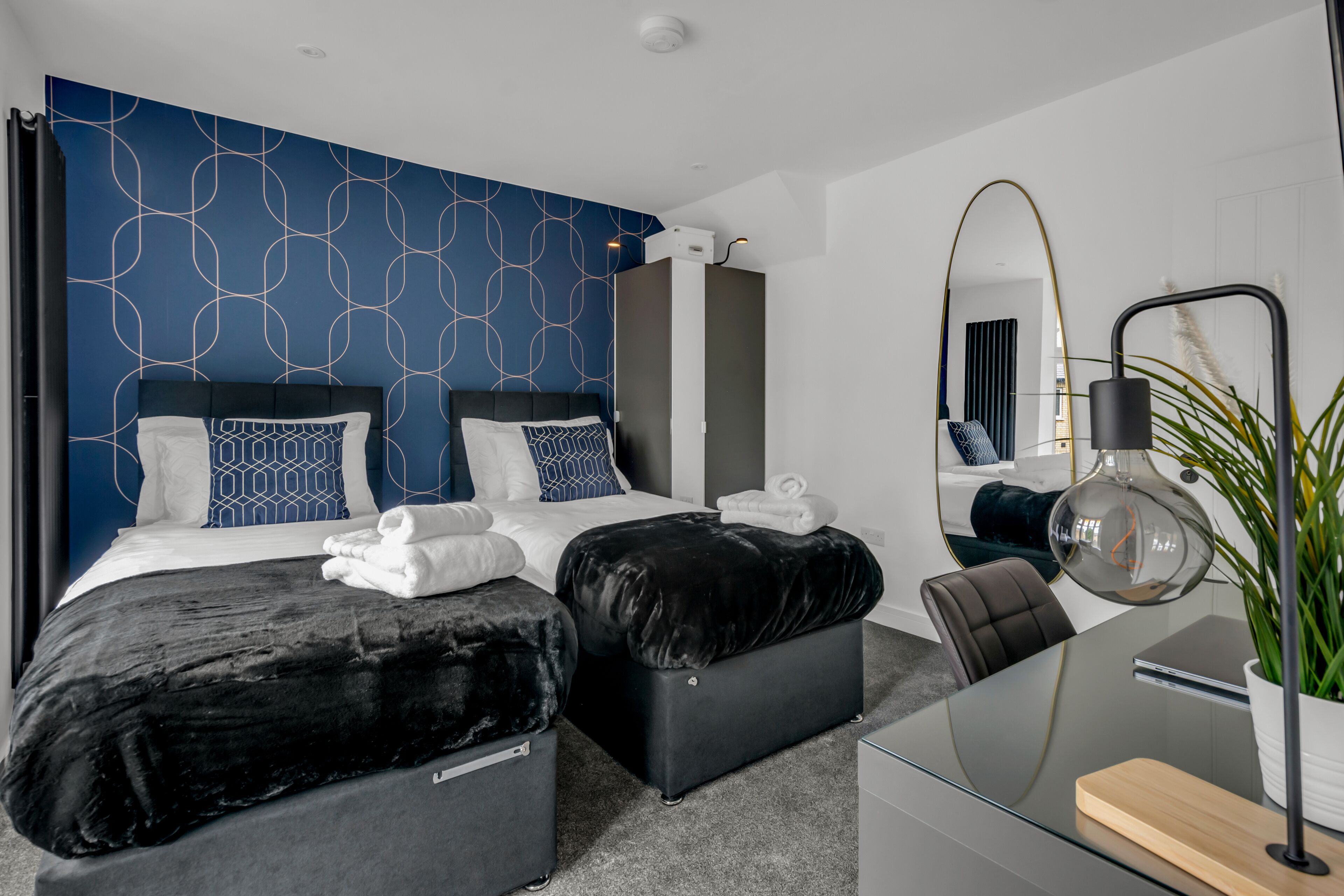 Ein modernes Hotelzimmer mit einem großen Bett, zwei schwarzen Hocker, einem Glastisch und einem Spiegel.