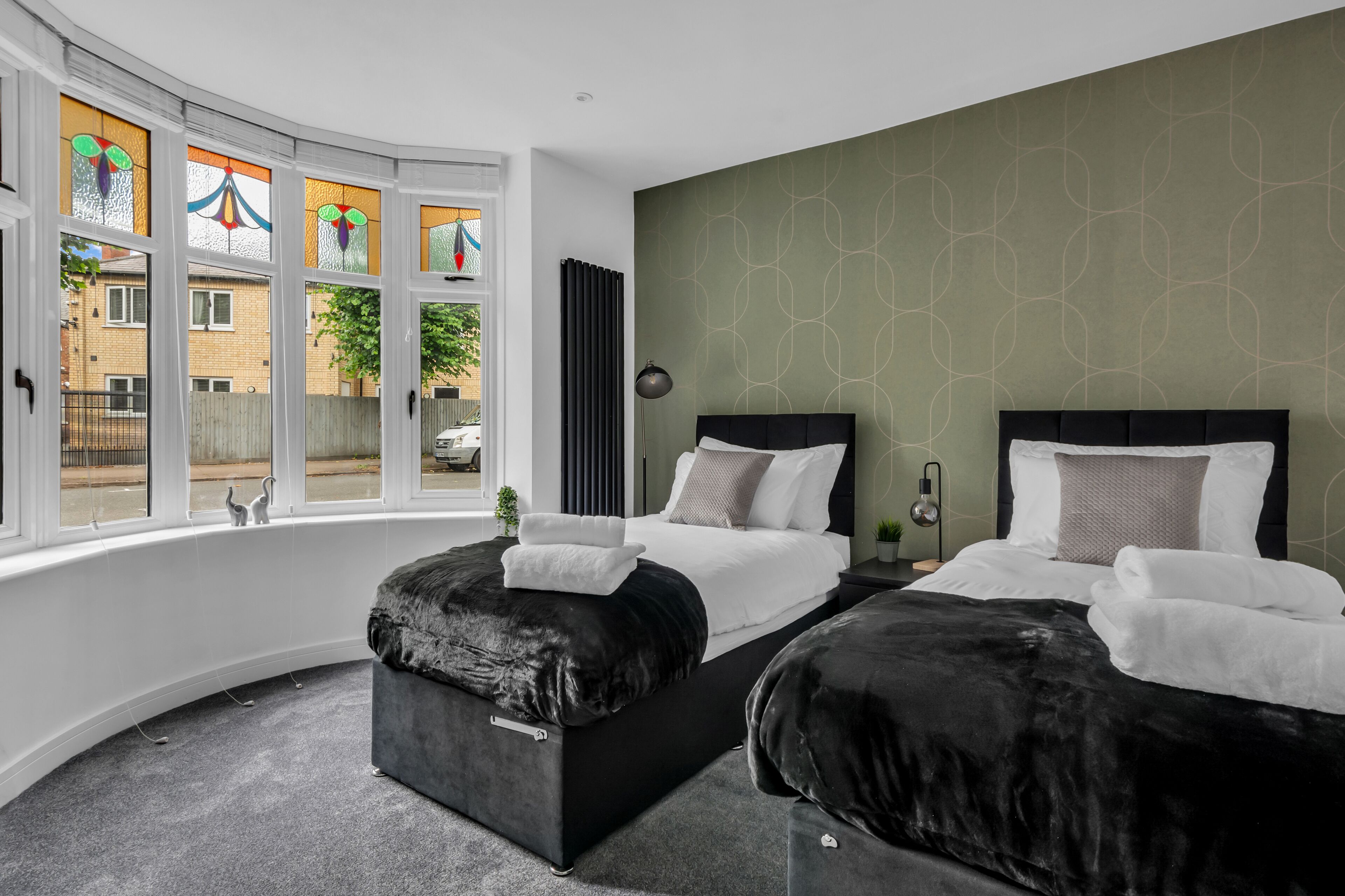 Quarto de hotel com duas camas, janela curva com vitrais, parede com padrão verde e vista para a rua.