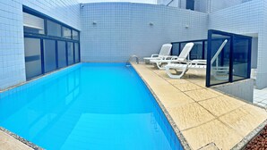A swimming pool with tiled walls and a tiled deck. There are white lounge chairs and a glass-enclosed area. - Vacanze - Infiniti (Maceió)