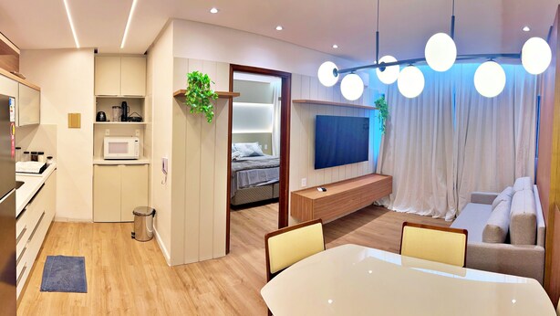 A modern hotel room with a kitchenette, a bed with a view, a TV, a seating area, and a dining table. - Vacanze - Infiniti (Maceió)