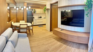 A modern living room with a curved wooden console, a flat-screen TV, a grey sofa, and a dining area with a white table and chairs. - Vacanze - Infiniti (Maceió)