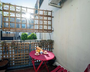 A red table with a plate of food, a cup, and a bottle, set against a white wall with a wooden fence and a window in the background. - Palais Hispania – 2P pop et coloré, face gare Nice (Nice)
