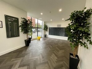 Un hall d’entrée spacieux avec un sol décoré d’un motif en chevrons, un grand panneau noir indiquant les numéros des chambres et des plantes en pot.