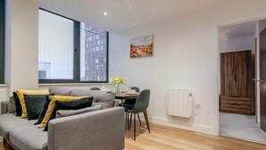 A modern living room with a grey sofa, a dining table with chairs, a radiator, and a large window with blinds. - 2-Bed | Old Trafford | Free Parking | Contractors (Stretford)