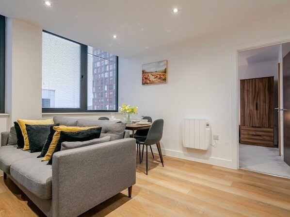A modern living room with a grey sofa, a dining table with chairs, a radiator, and a large window with blinds.