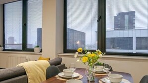 A dining area with a table set for four, two potted plants on the windowsill, and a cityscape view through the windows. - 2-Bed | Old Trafford | Free Parking | Contractors (Stretford)