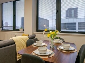 A dining area with a table set for four, two potted plants on the windowsill, and a cityscape view through the windows.