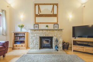 A living room with a stone fireplace, a wooden mantel, a television on a wooden stand, and a bookshelf. - Stunning Cotswolds Barn Conversion | Spacious for Groups & Families (Kempsford)