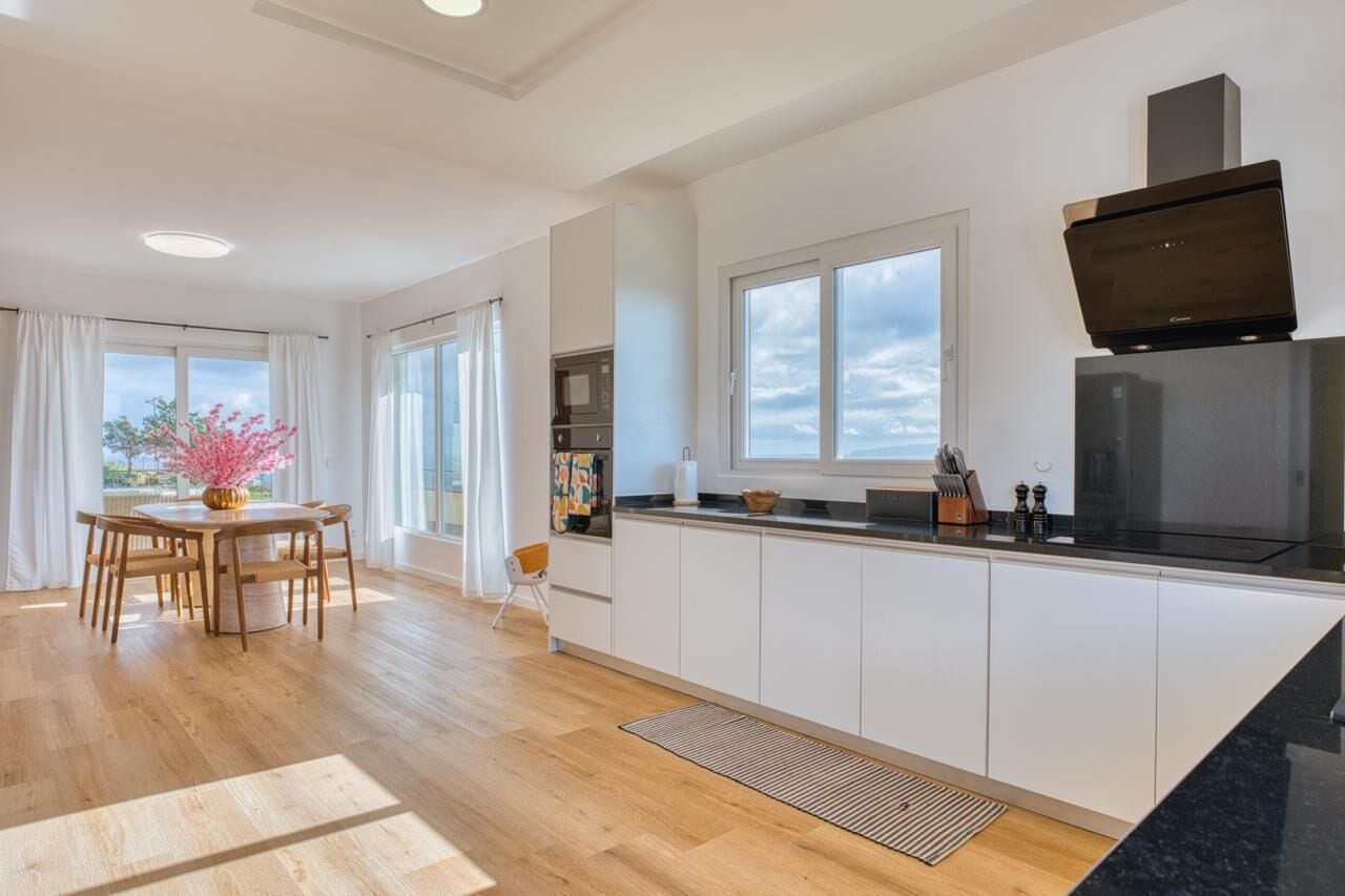 A modern kitchen with white cabinetry, a black countertop, and a built-in oven. There is a dining area with a wooden table and chairs, and a window with a view of the sky.