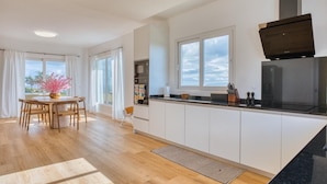Une cuisine moderne avec des meubles blancs, un plan de travail noir et un four encastré. On y trouve un coin repas avec une table et des chaises en bois, ainsi qu’une fenêtre donnant sur le ciel.
