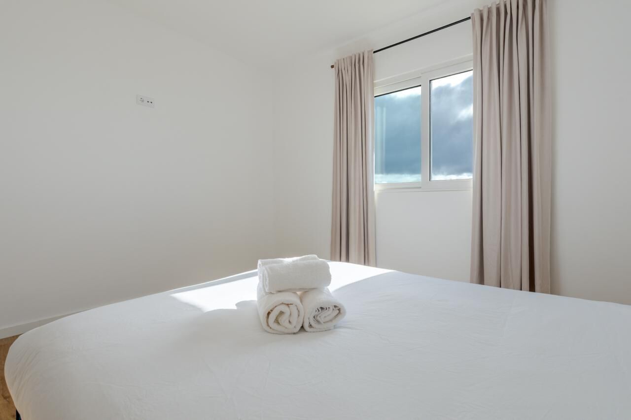 Een netjes opgemaakt bed met opgerolde handdoeken, een raam met gordijnen en een stopcontact aan de muur.