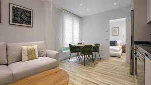 A modern living room with a sofa, coffee table, and dining area with chairs. There is a kitchen visible in the background. - Dobo Madrid Pinar Aptos Temporales (Madrid)
