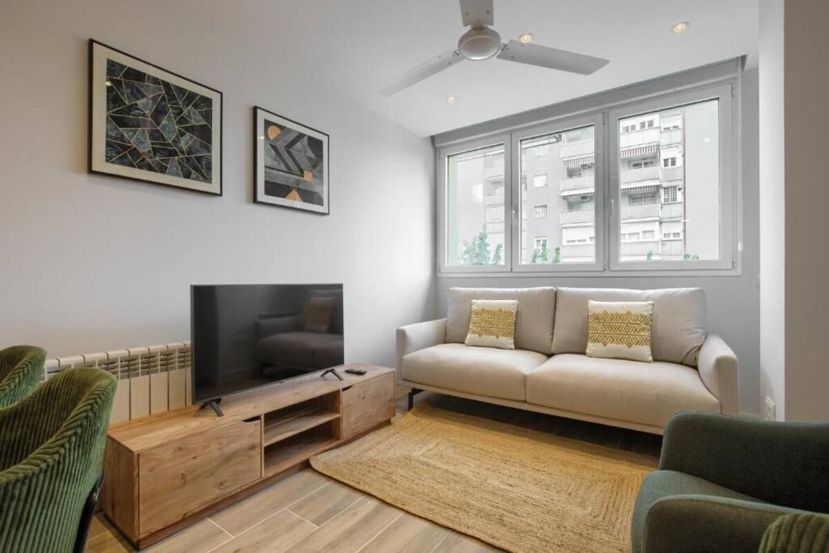 Ein modernes Wohnzimmer mit einem Flachbildfernseher auf einem Holzboden, einem beigefarbenen Sofa mit gemusterten Kissen und einem großen Fenster mit Blick auf die Stadt.