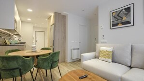A modern living room with a round wooden coffee table, a grey sofa with patterned cushions, and a dining area with green chairs and a wooden table. - Dobo Madrid Pinar Aptos Temporales (Madrid)