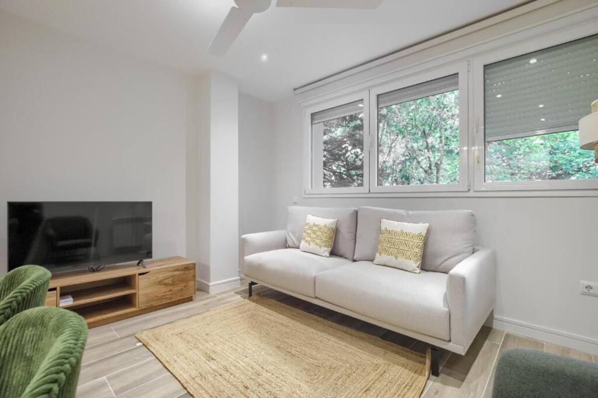 Ein modernes Wohnzimmer mit einem Flachbildfernseher, einem grauen Sofa mit gemusterten Kissen, einem Holz-TV-Schrank und einem großen Fenster mit Blick auf Bäume.