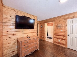 A wooden-paneled room with a flat-screen TV mounted on the wall, a wooden dresser with drawers, and a white door. - Pet-Friendly  Mountain Cabin | Hot Tub & Fire Pit (Sautee Nacoochee)