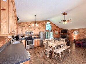 A spacious kitchen with wooden cabinetry, a dining area with a wooden table and benches, and a living room with a sofa and a television. - Pet-Friendly  Mountain Cabin | Hot Tub & Fire Pit (Sautee Nacoochee)