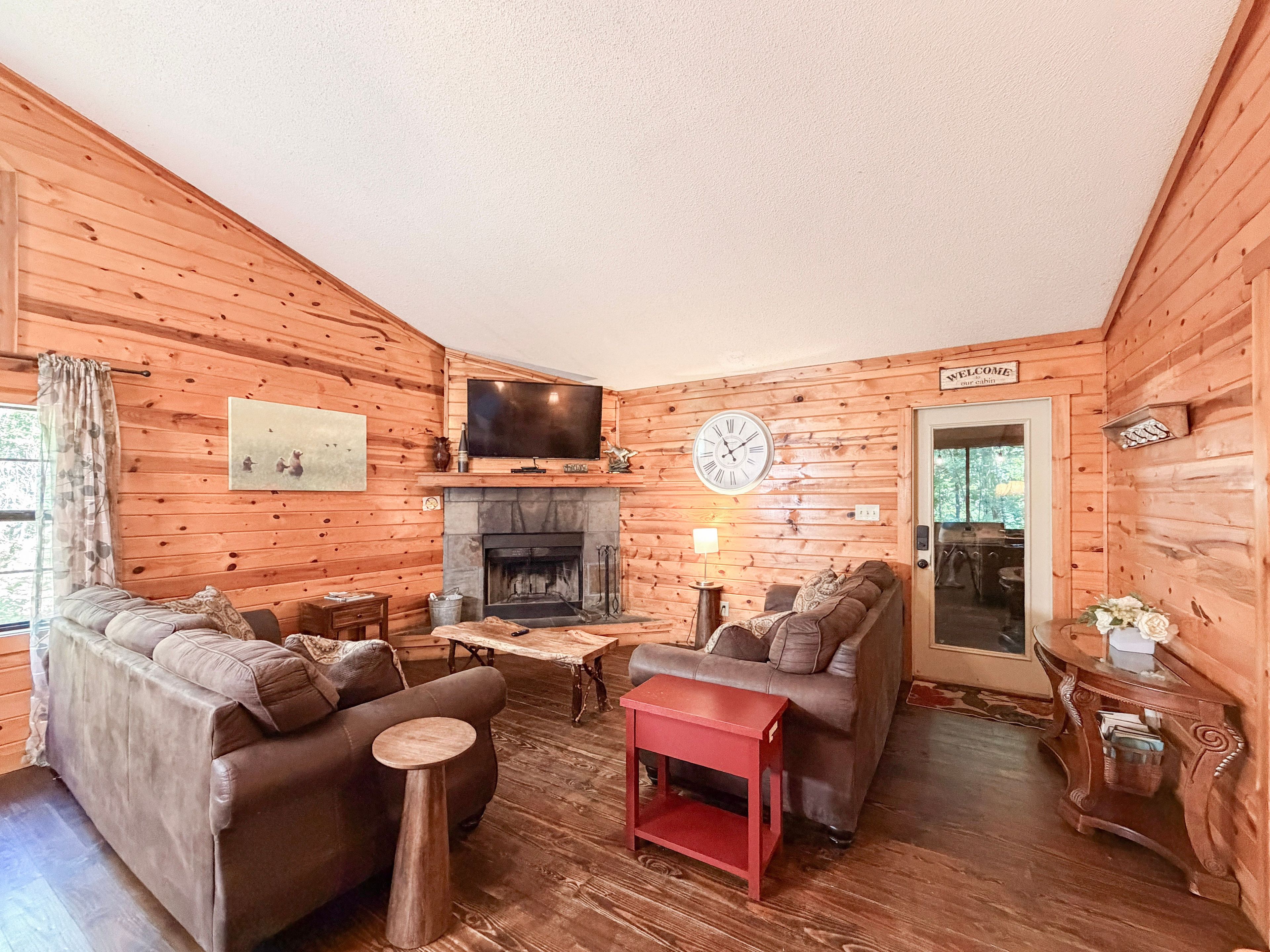 A cozy cabin interior with a fireplace, two sofas, a television, and a clock.