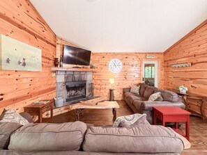 Ein Interieur einer Blockhütte mit Kamin, einem darüber montierten Fernseher, einer Couch, einem roten Tisch und einer Wanduhr.