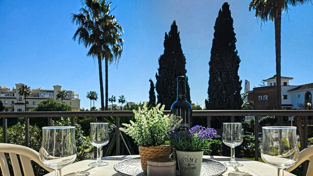 Una mesa dispuesta con copas de vino, una botella y plantas en macetas en un balcón con vistas a árboles altos y edificios.