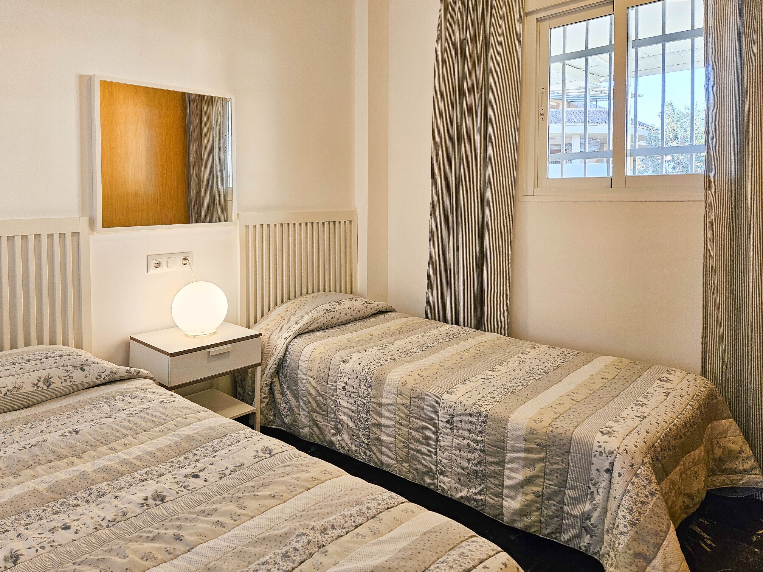 A bedroom with two beds, a bedside table with a lamp, a window with curtains, and a radiator.