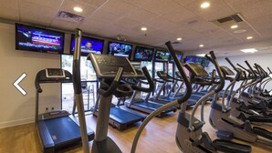 Ein Fitnessraum mit mehreren Laufbändern und Ergometer, einem an der Wand montierten Fernseher und Holzboden.