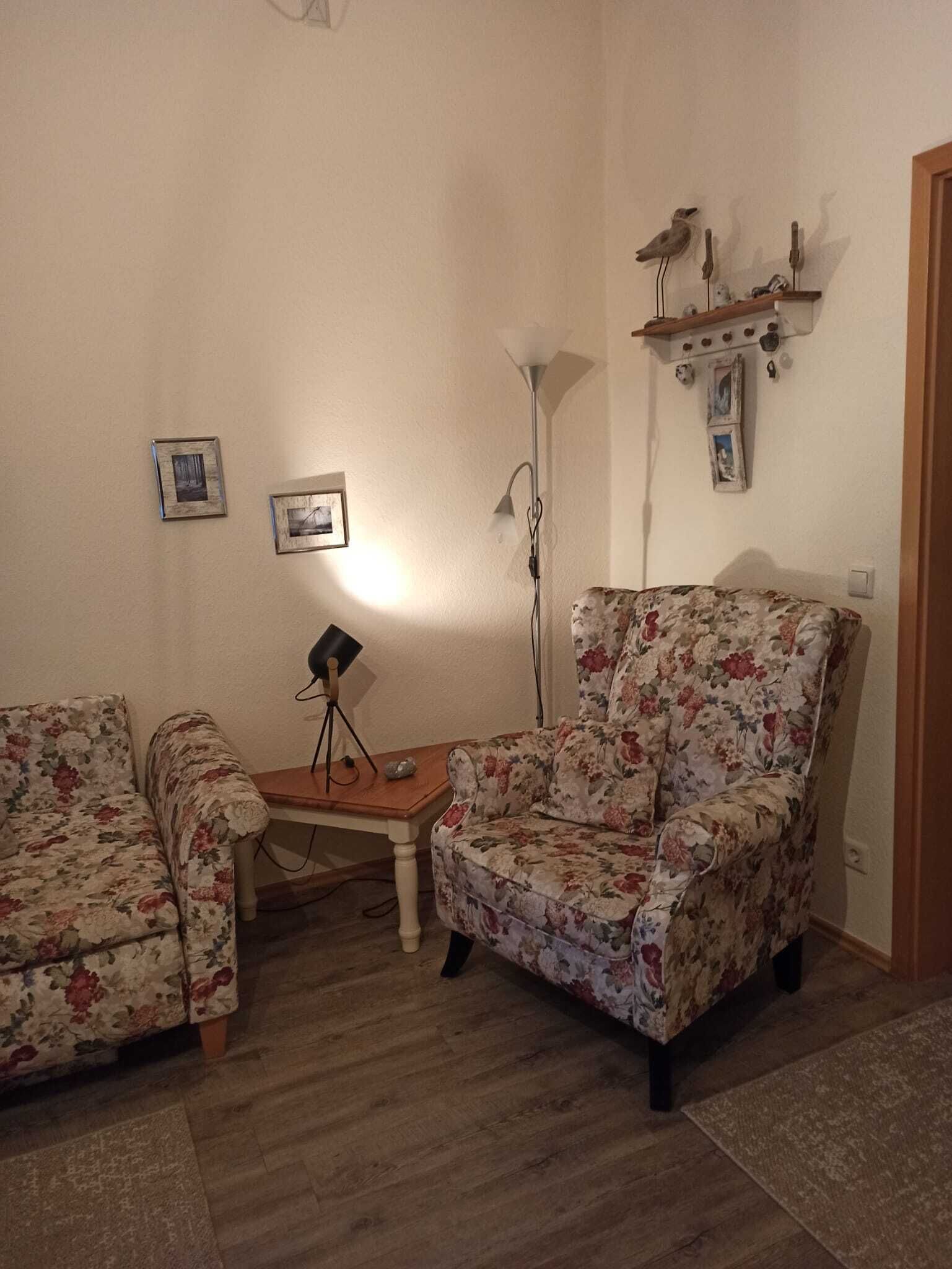 Ein Wohnzimmer mit zwei floralen Sesseln, einem Holztisch, einer Stehlampe und Wandregalen mit Dekoration.
