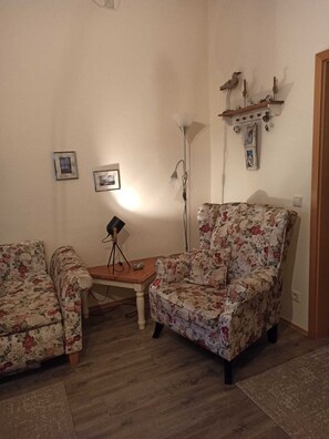 Ein Wohnzimmer mit zwei floralen Sesseln, einem Holztisch, einer Stehlampe und Wandregalen mit Dekoration.