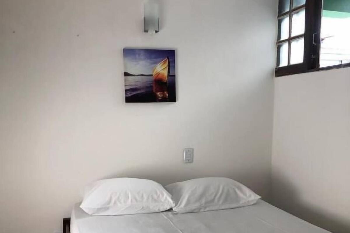 A single bed in a room with white walls, a painting of a sailboat on the wall, and a window with blinds.