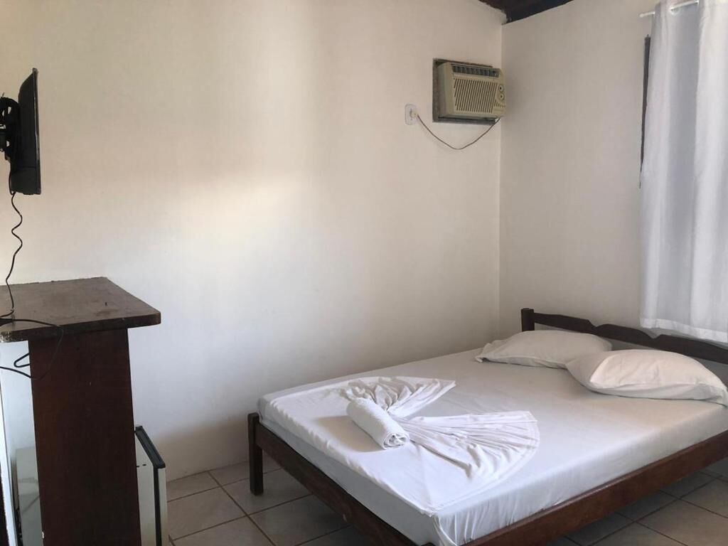 Una cama individual con ropa blanca y una toalla, un escritorio pequeño con una silla, un calentador fijado a la pared y una ventana con cortinas.