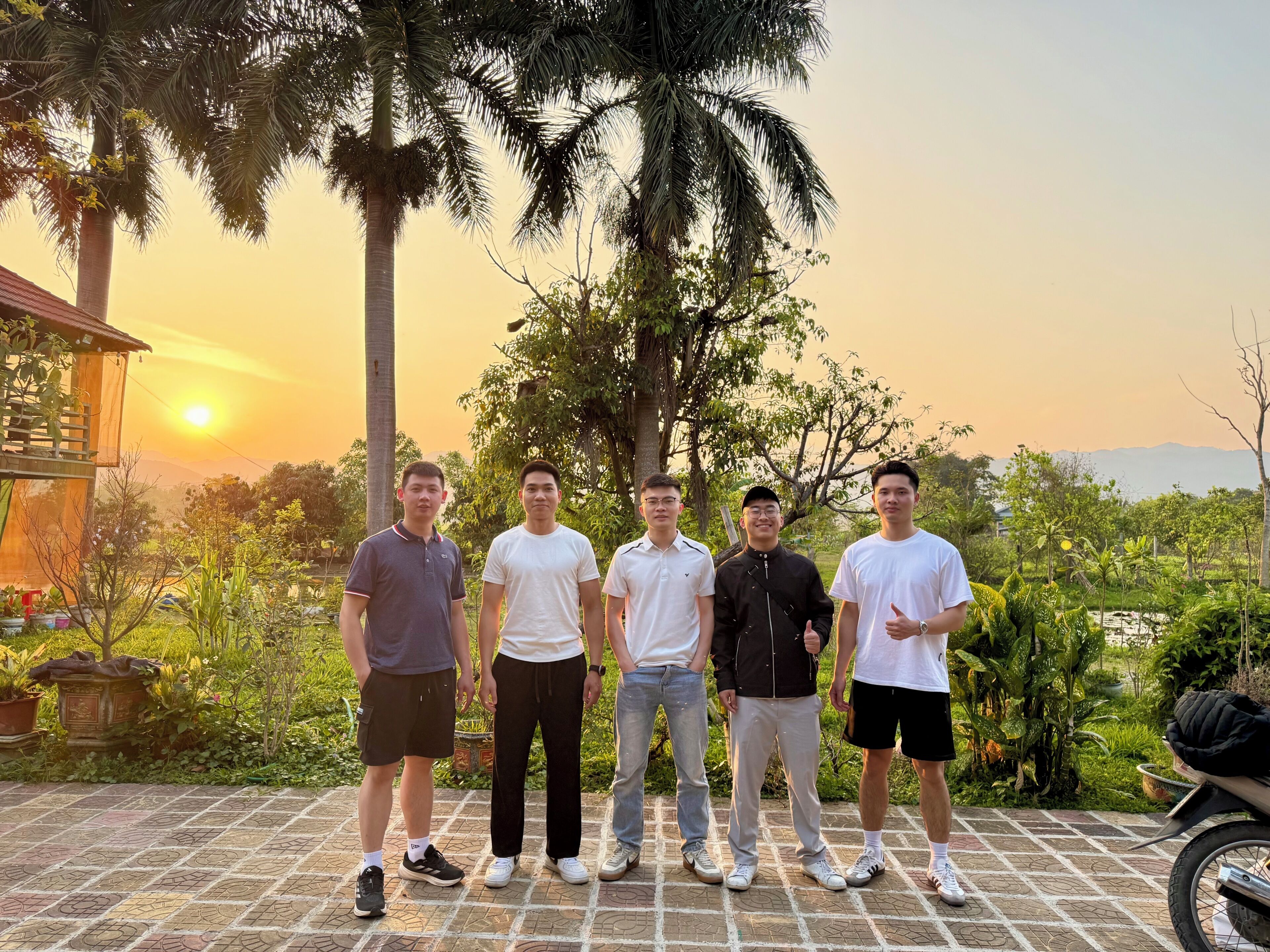 Un groupe de personnes se tient sur une surface pavée, avec un coucher de soleil en arrière-plan.