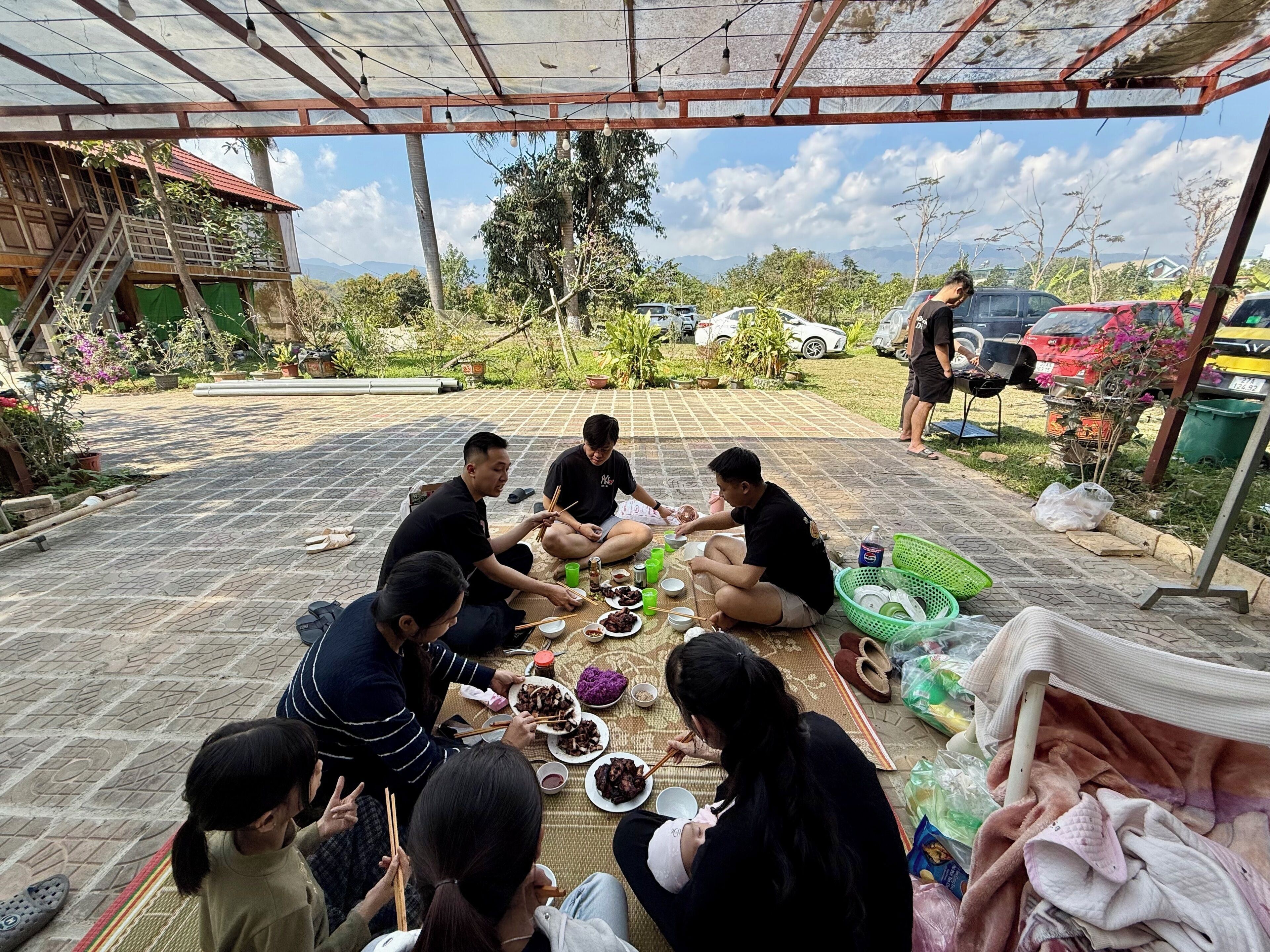 A group of people sitting on a patterned mat outdoors, eating with chopsticks.