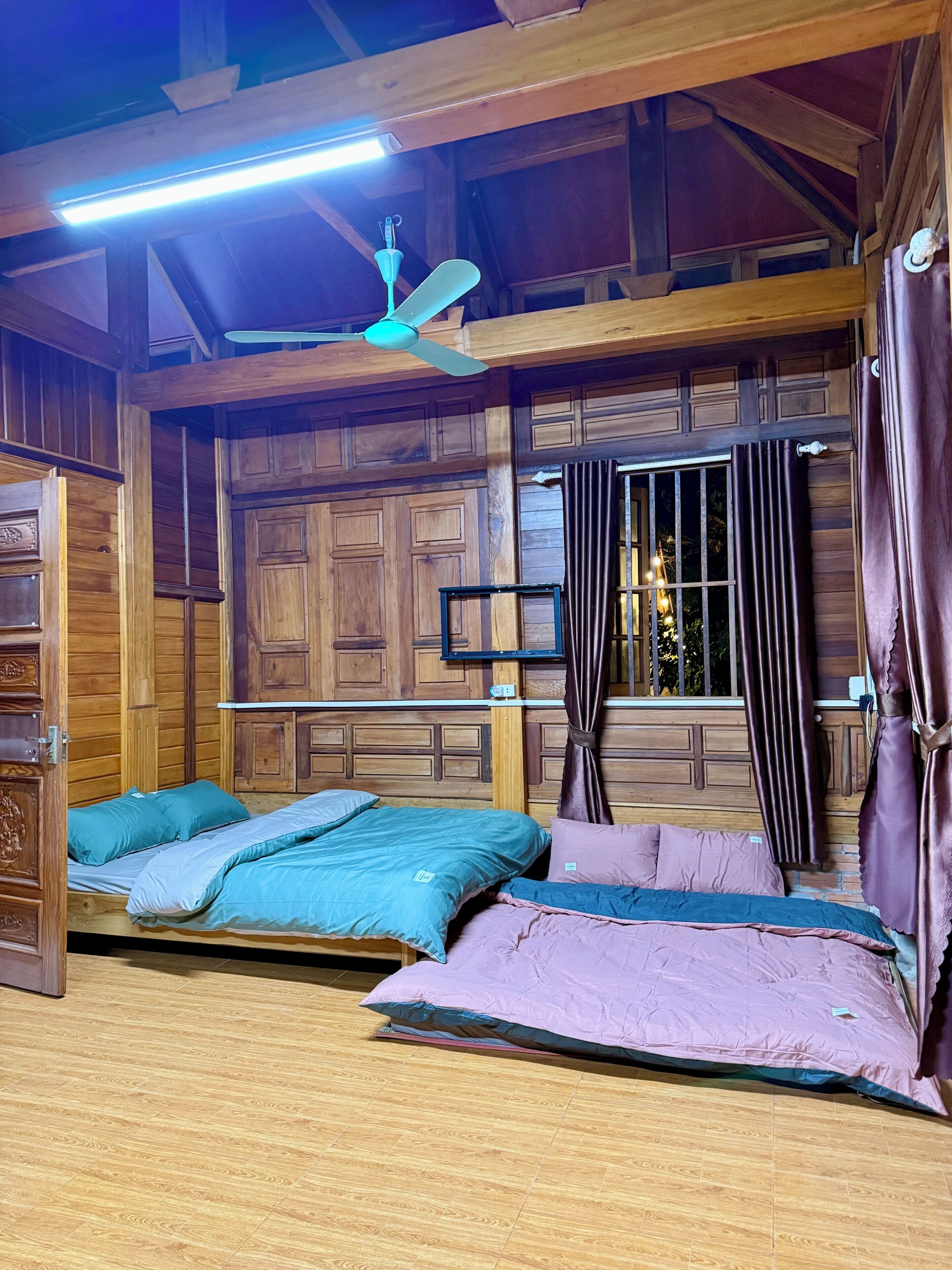 Una habitación con paneles de madera, ventilador de techo, cama con ropa de cama azul y rosa, y ventana con cortinas.