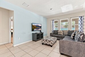 A living room with a flat-screen TV, a grey sofa, patterned cushions, and a patterned ottoman. - 25% Off 5BR Retreat! Pool/Movie Room! (Davenport)