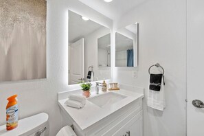 Une salle de bain moderne avec un lavabo blanc, un miroir et des toilettes. On y trouve un porte-serviettes avec une serviette noire et un flacon de désodorisant sur le comptoir.