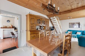 A wooden dining table with chairs, a kitchen area with cabinets, a staircase, and a blue sofa in a room with wooden ceiling beams. - Gite de la Closerie – Independent Country House, Perfect for Families (Oisly)