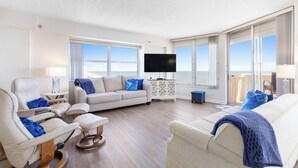 A spacious living room with a large window, a flat-screen TV, and a comfortable seating area with a sofa and chairs. - New listing! 3-bedroom. Ocean-Front, Corner Unit with Panoramic Views (Ormond Beach)