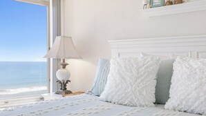 A bedroom with a large window, a bed with white and light blue pillows, and a lamp on a bedside table. - New listing! 3-bedroom. Ocean-Front, Corner Unit with Panoramic Views (Ormond Beach)