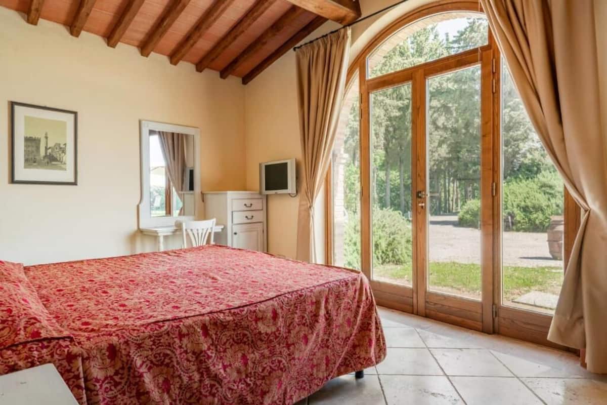 A bedroom with a bed, a desk, a chair, a television, and a large window with curtains.