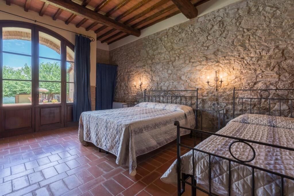 A bedroom with two beds, stone walls, a wooden ceiling, and a view through a window.