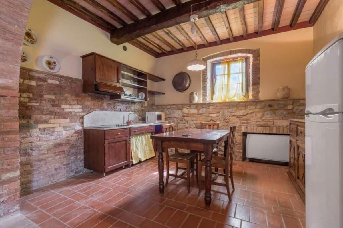 A rustic kitchen with exposed brick walls, a wooden table, and a stone fireplace.