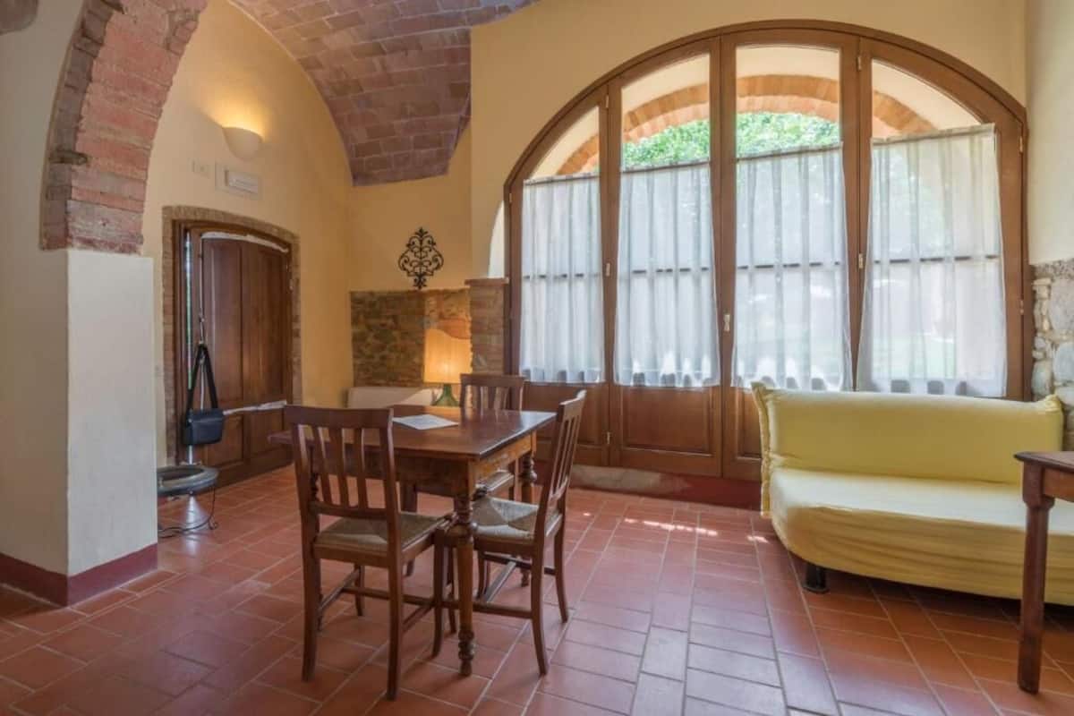 A dining area with a wooden table and chairs, a yellow sofa, and a large arched window with curtains.