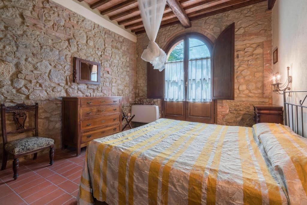 Ein Schlafzimmer mit Steinwand, einem Bett mit gestreifter Bettdecke, einer hölzernen Kommode, einer Bank und einem Fenster mit Vorhängen.