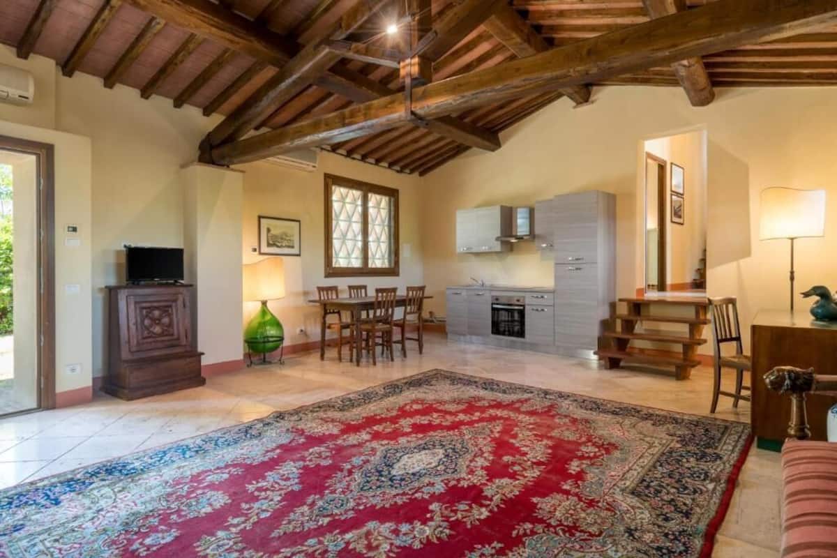 A spacious living area with a large rug, wooden ceiling beams, a dining table, and a kitchen area.