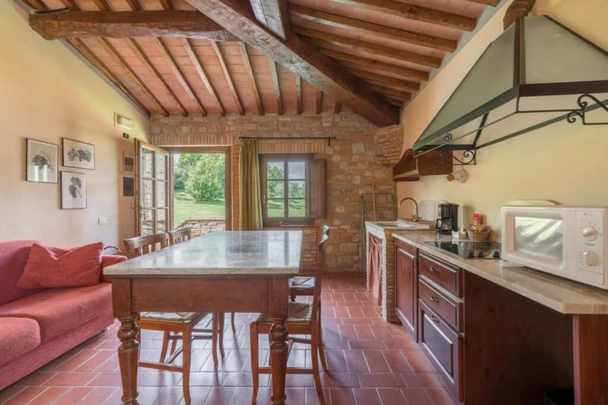 A rustic kitchen with a stone wall, wooden beams, a dining table, and a red sofa.