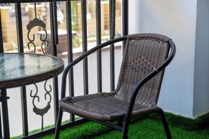 A wicker chair with a curved backrest and a round table with a glass top, positioned on a balcony with a metal railing and a view of a building in the background. - Nested in the Heart of Nairobi, Modern furnished with a safe for your valuables. (Nairobi)