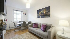 A living room with a sofa, a dining table with chairs, a TV, and a painting of a bridge. - Welcoming 1-bedroom apartment in the heart of London (London)