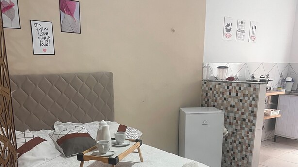 Ein ordentlich bezogenes Bett mit weißer Bettdecke, ein Nachttisch mit Tasse und Untertasse sowie ein kleiner Kühlschrank.