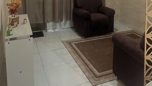 Ein Zimmer mit einem braunen Sessel, einem weißen Tisch mit einer Vase und einer Flasche, einem gemusterten Teppich und einem Fenster mit durchsichtigen Vorhängen.